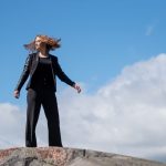 Young woman posing confidently on a rock in a Helsinki park, with a clear blue sky and European architecture in the background.